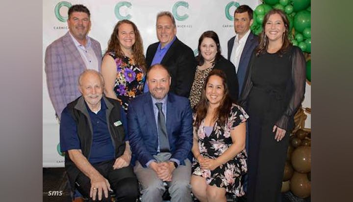 A group of nine people posed together, dressed in formal attire, in front of a logo backdrop and green balloon decorations.
