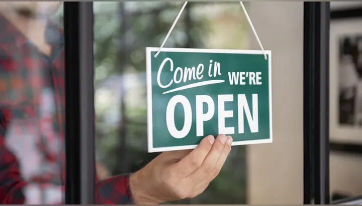Hand placing a green "Come in, we're open" sign on a glass door.