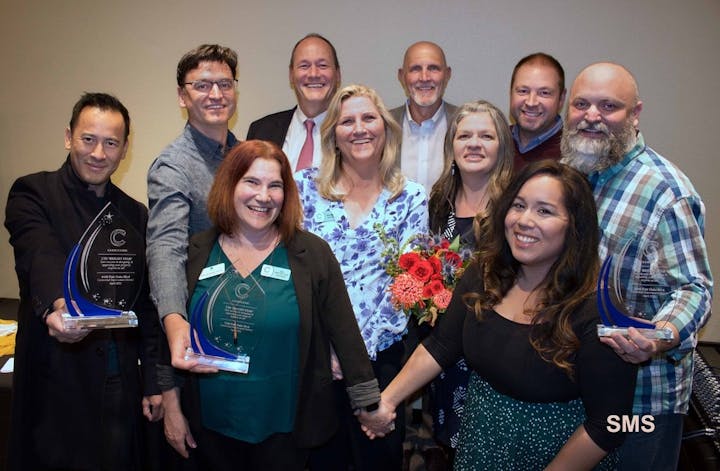 A group of people smiling, holding awards and flowers at an event.