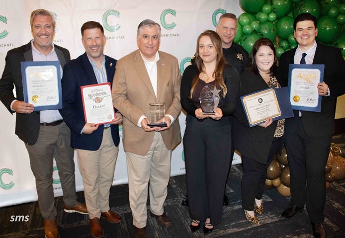 A group of people posing with awards and certificates in front of a backdrop with "Carmichael" and green balloons.