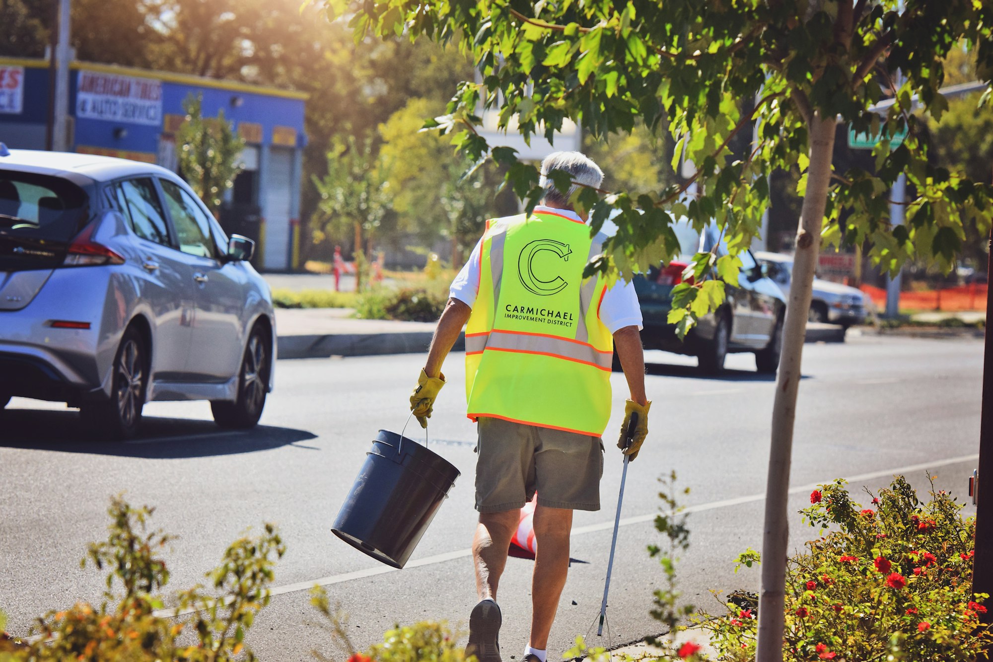 A man in a neon vest cleans a street, carrying a bucket and a grabber tool. Cars drive by, with trees and flowers along the sidewalk.