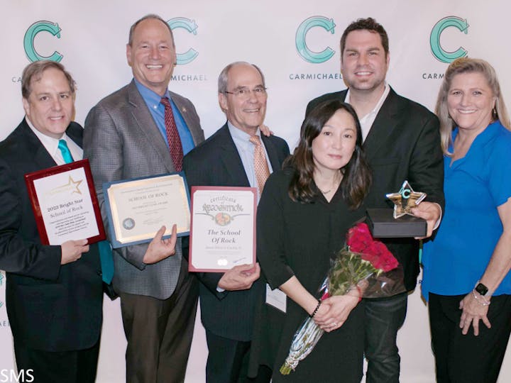 Group of people at an event, holding awards and certificates from "The School of Rock."