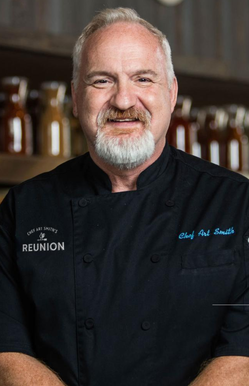 A smiling man in chef's attire with "REUNION" on the jacket in a kitchen-like setting.