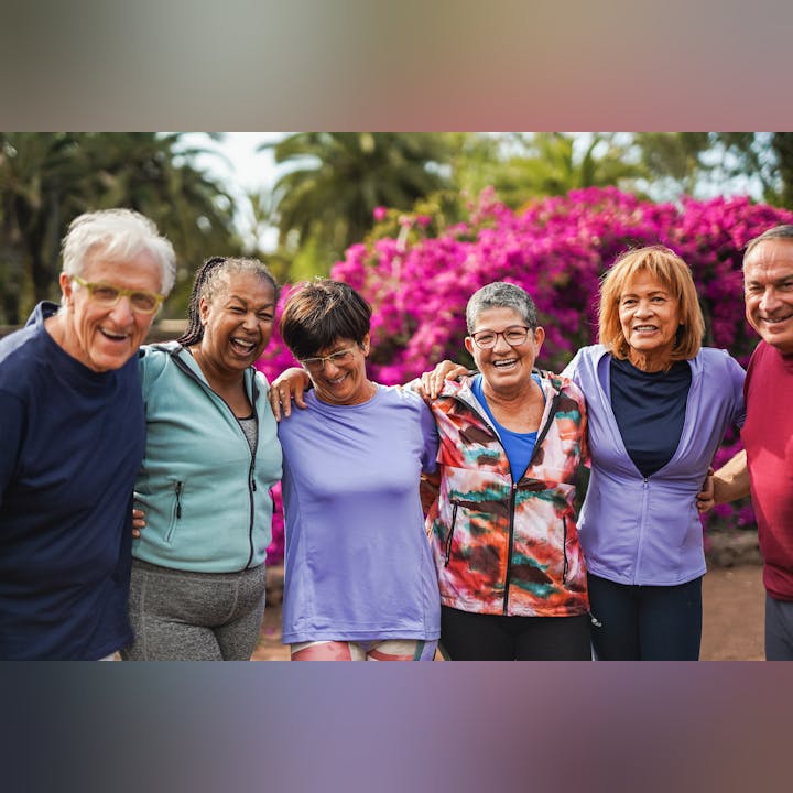 A group of six smiling people with arms around each other, standing outdoors in front of vibrant pink flowers.