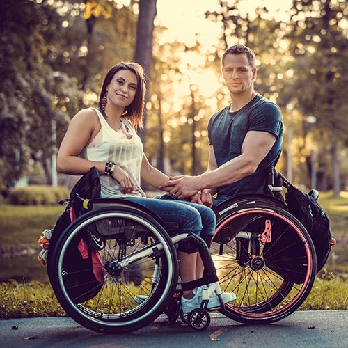 Two people in wheelchairs holding hands in a park at sunset.