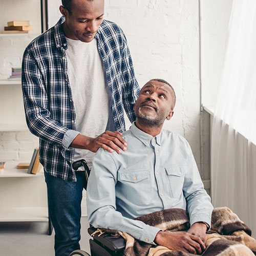A standing man comforting a seated older man who looks up at him, both indoors.