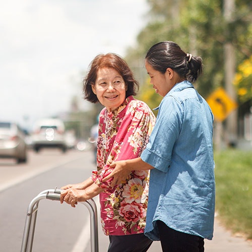 A younger woman assists an elderly lady using a walker by the roadside.