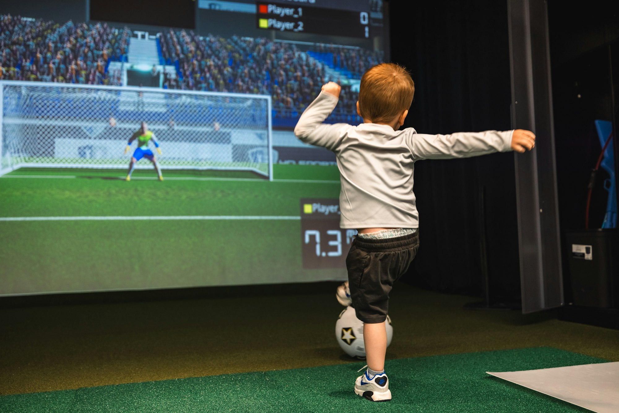 A child imitates kicking a soccer ball towards a virtual goal on a screen, with scoring graphics displayed above.