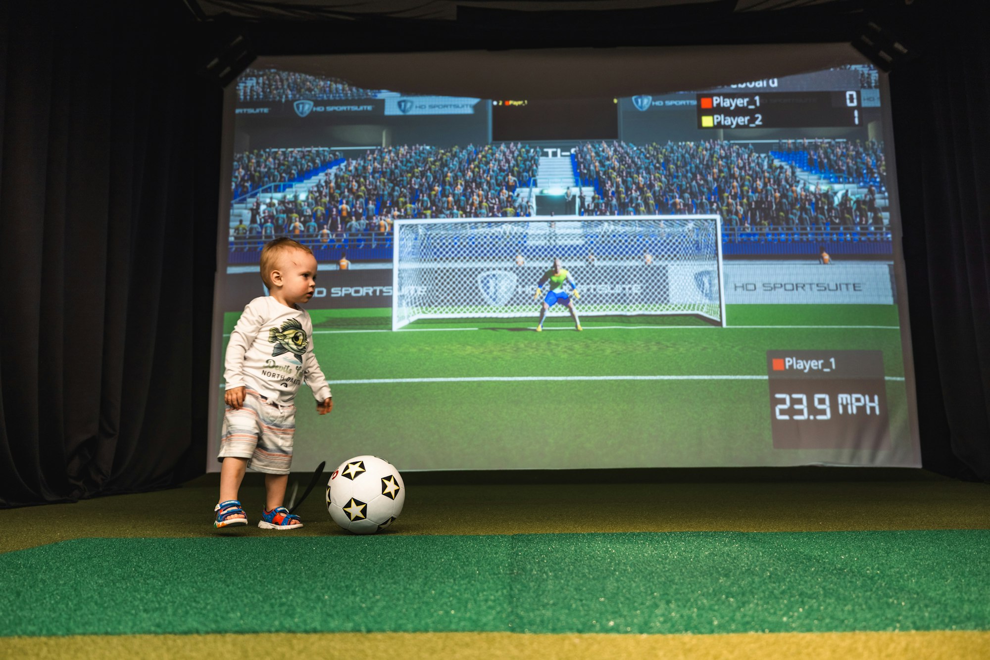 A toddler stands near a soccer ball with a virtual soccer game screen in the background.
