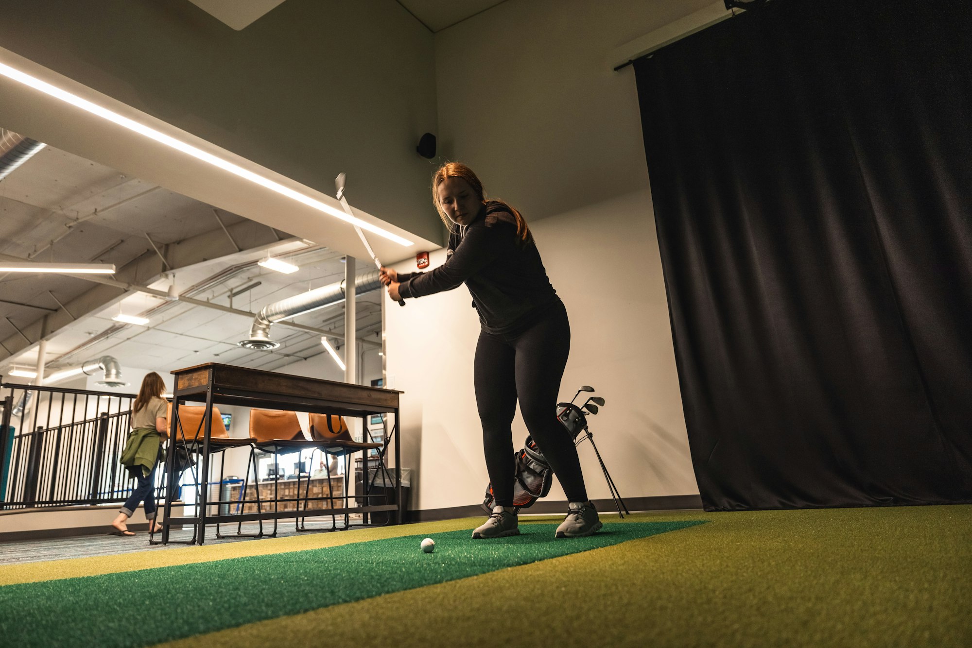 A woman is indoor golfing; a golf bag stands nearby, another person in the background. Modern, industrial-style interior.
