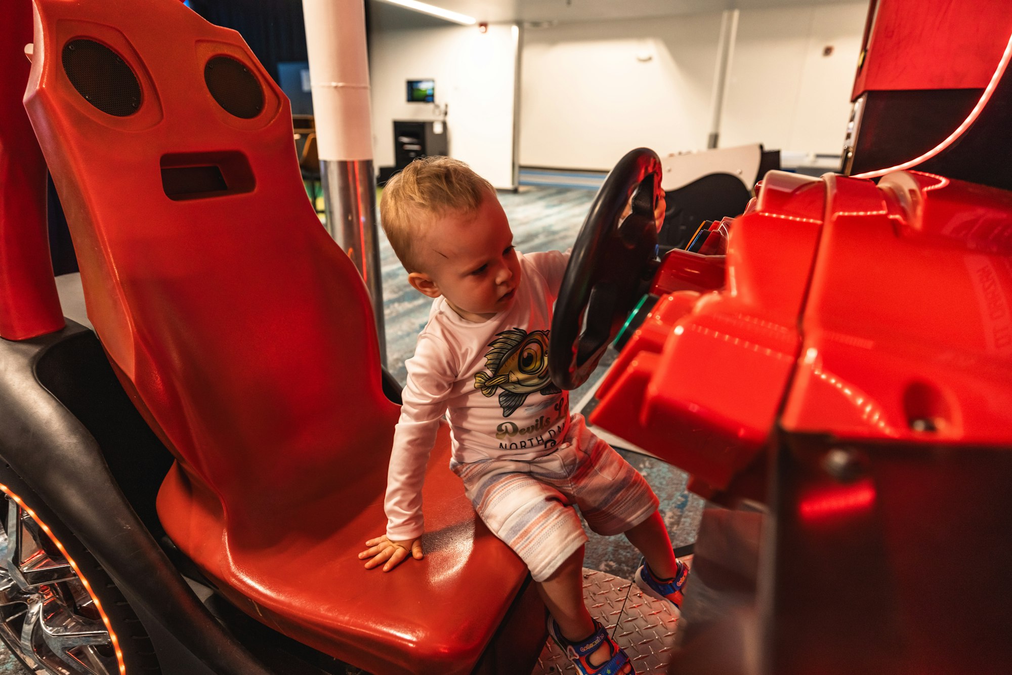 A toddler playing with a red arcade racing game steering wheel.