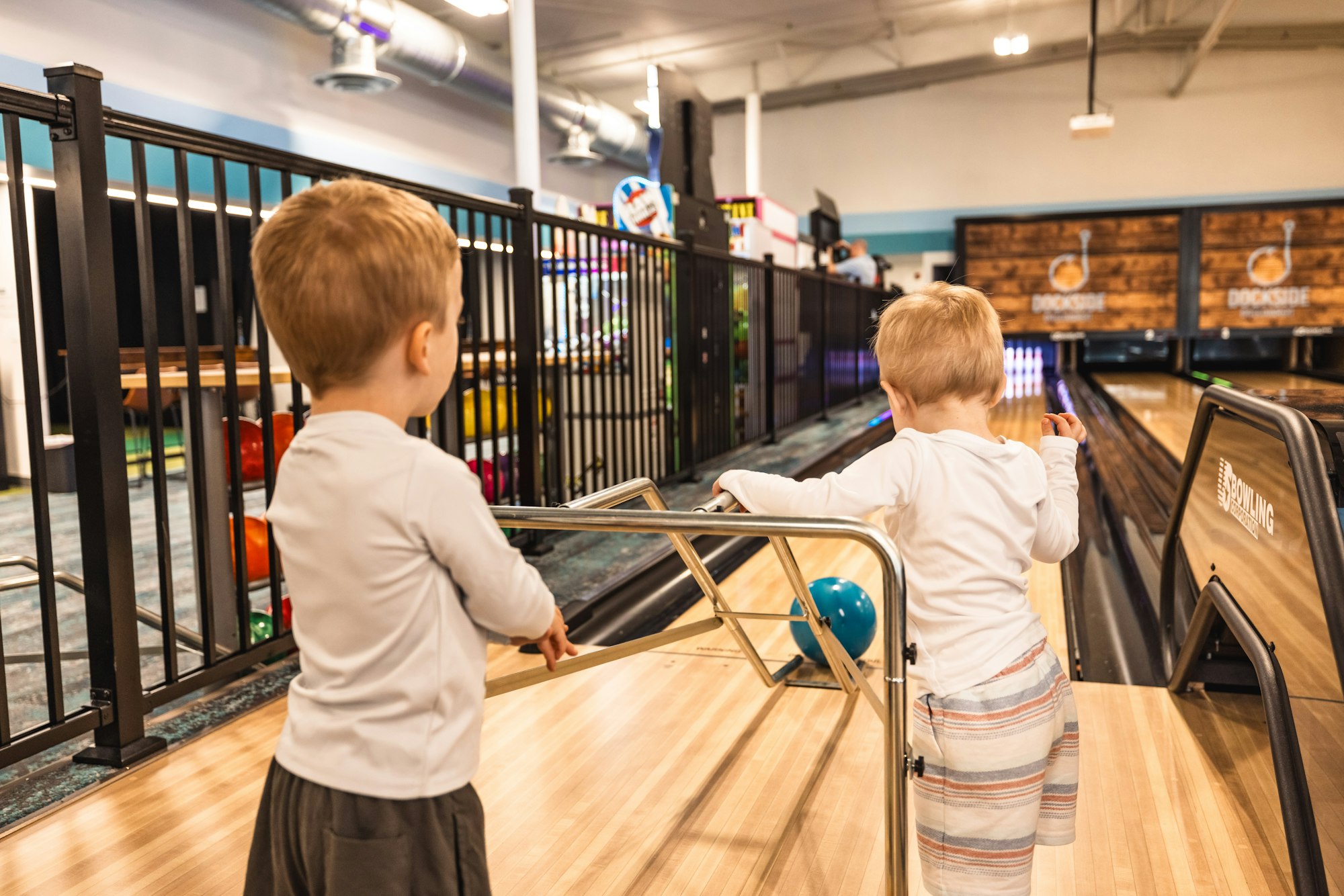 Two kids are watching a bowling lane, with one kid seemingly about to roll a ball down the alley.