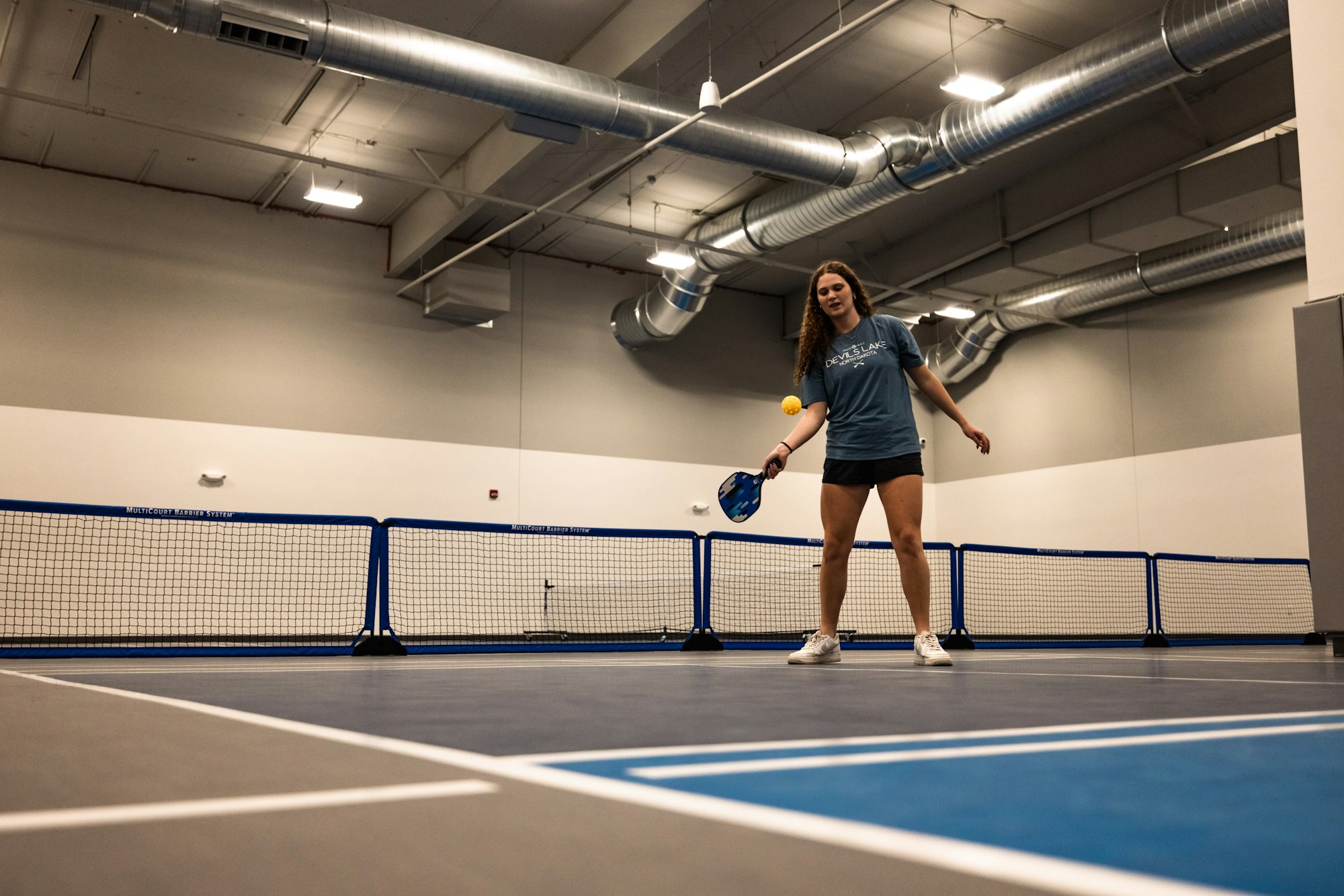 A person playing pickleball indoors, equipped with a paddle and a ball, standing by the net.