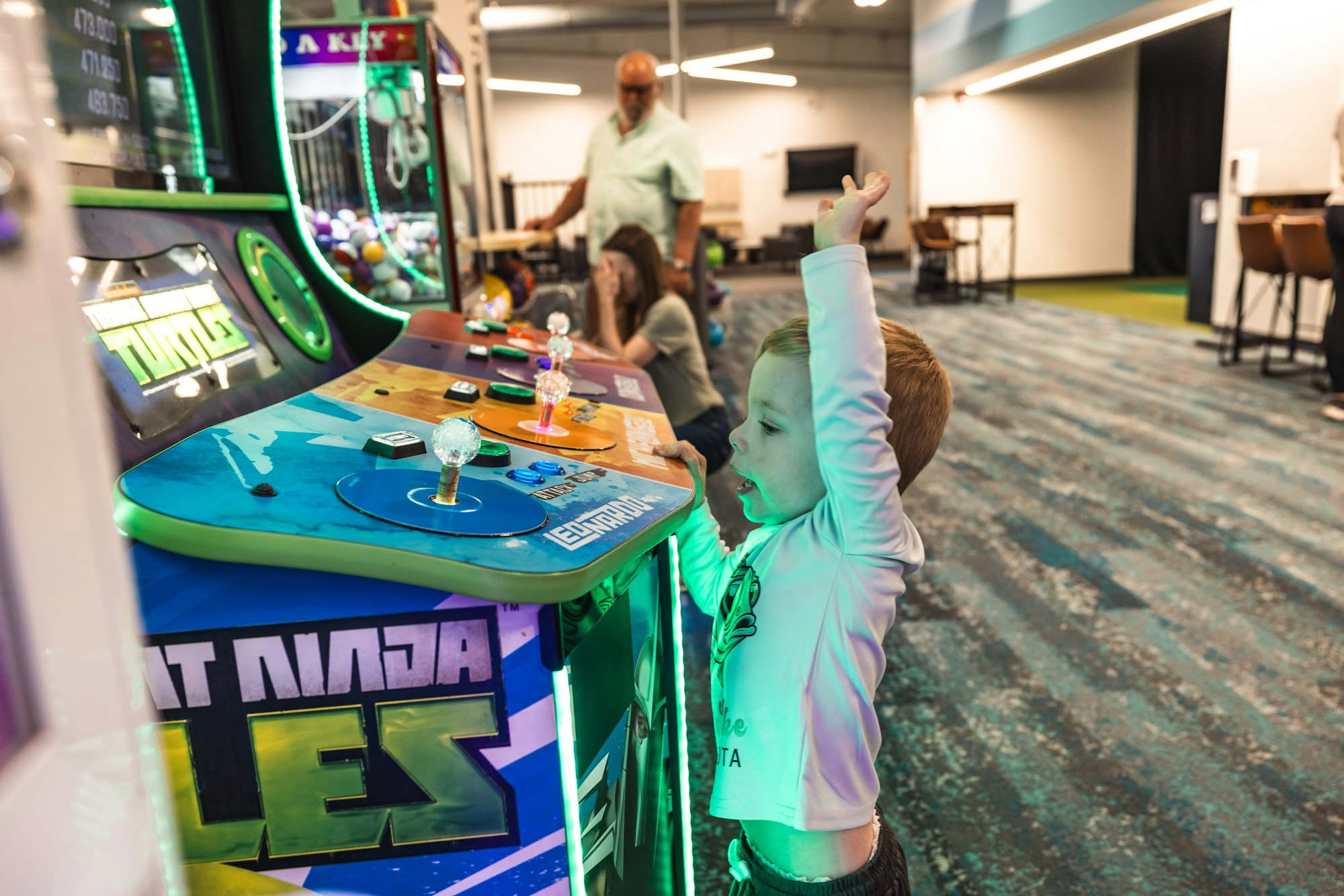 Child playing in an arcade with a joystick, excitedly raising hand, adult supervising in background.