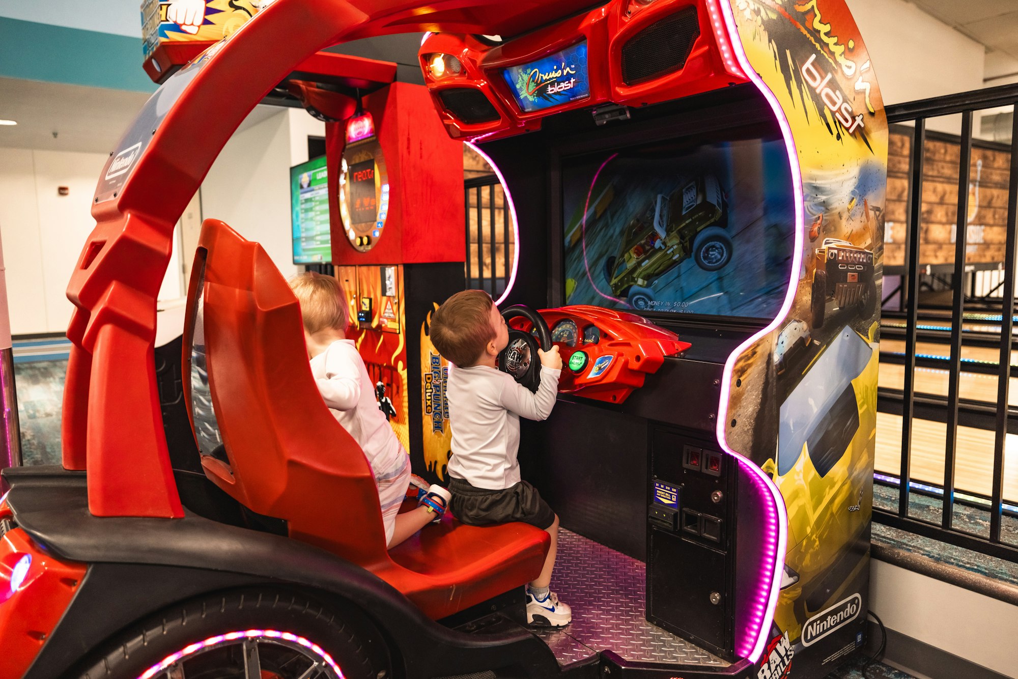 Two kids playing an arcade racing game, "Cruis'n Blast," with bright lights and colorful graphics.