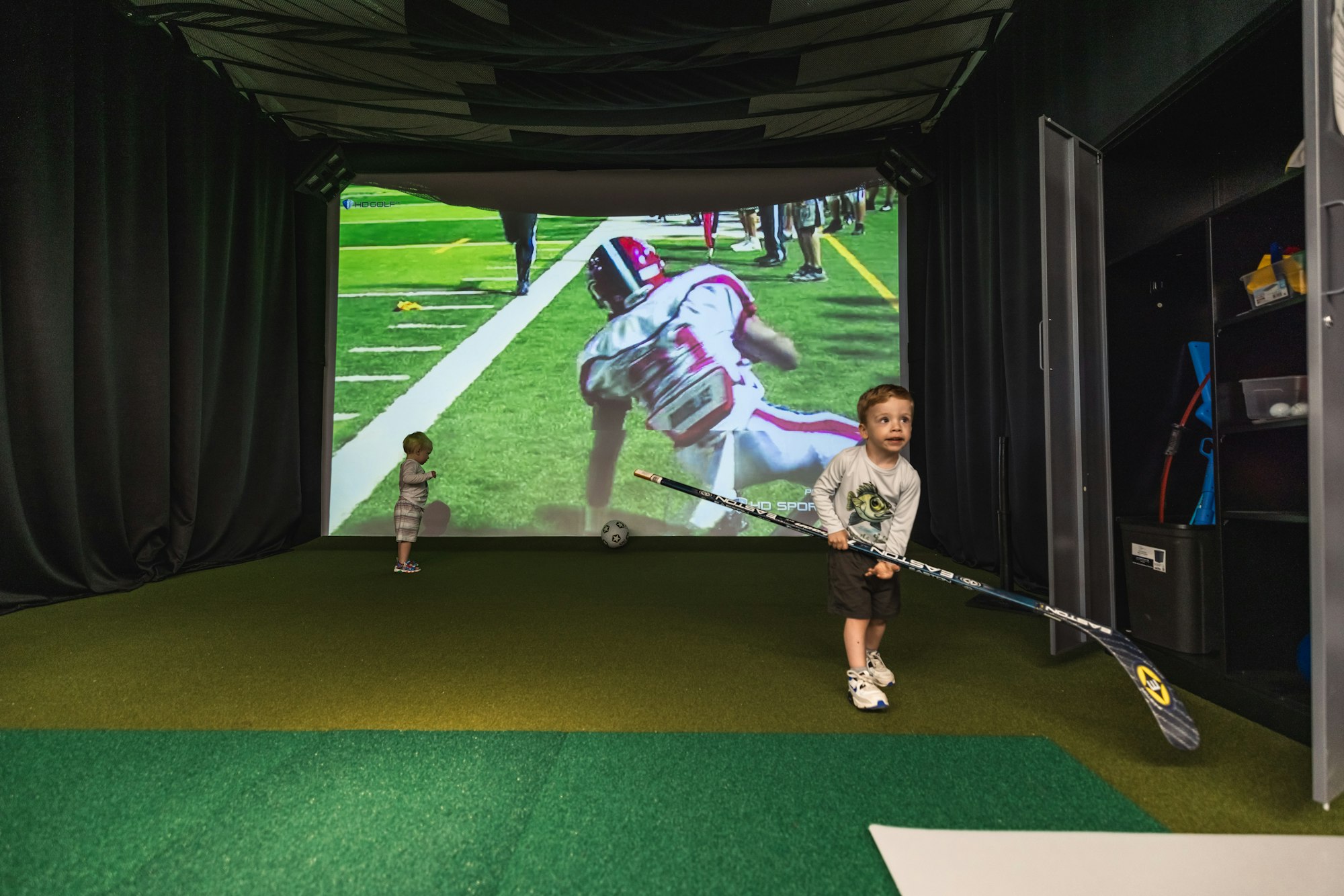 Two kids standing in a simulation room with a large screen displaying a sports game, artificial turf floor, and sports equipment around.