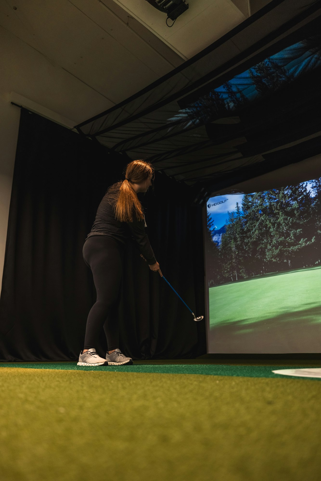A person is using a golf club in an indoor golf simulator with a large screen displaying a virtual course.