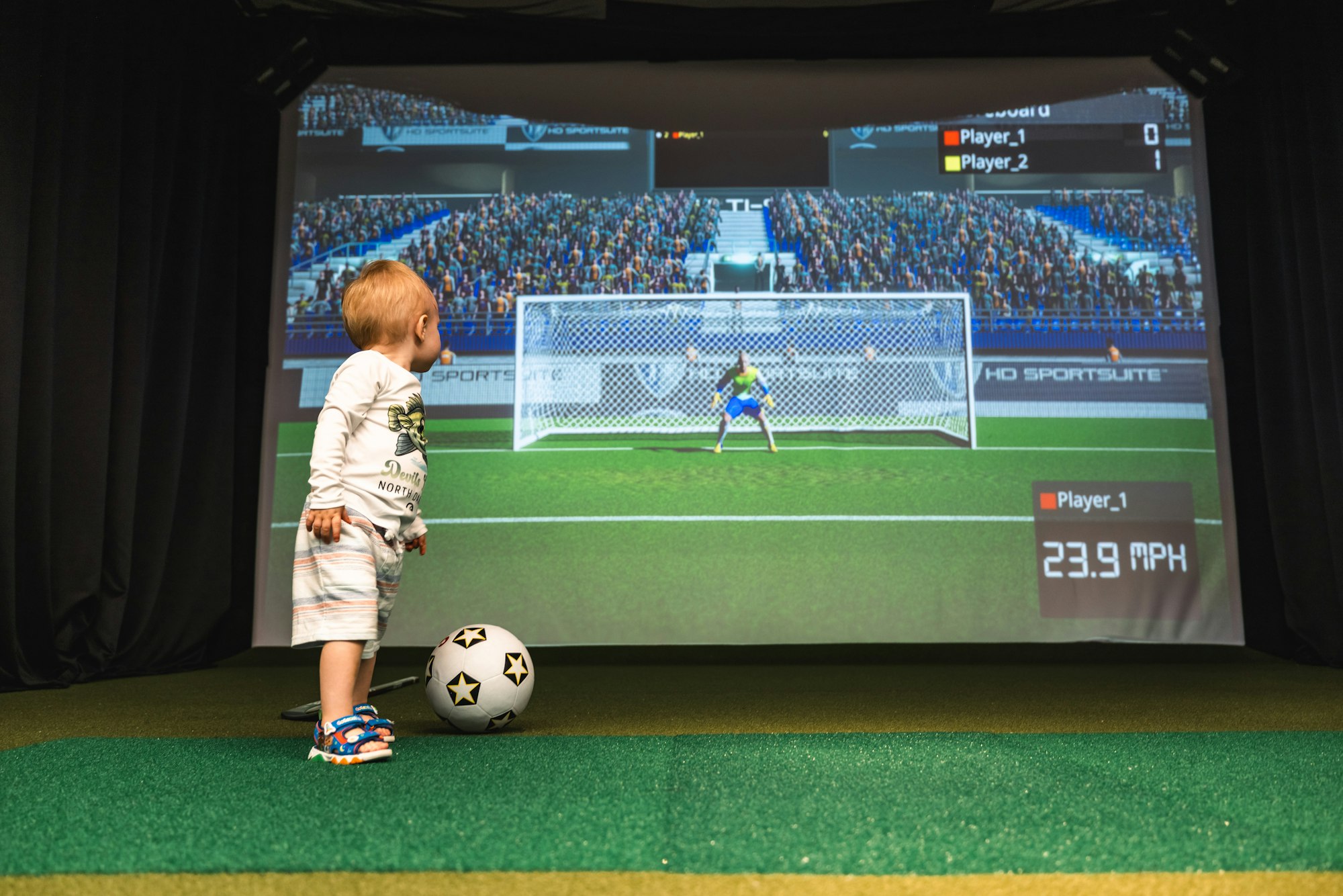 A child playing with a soccer ball in front of a large screen simulating a soccer game.
