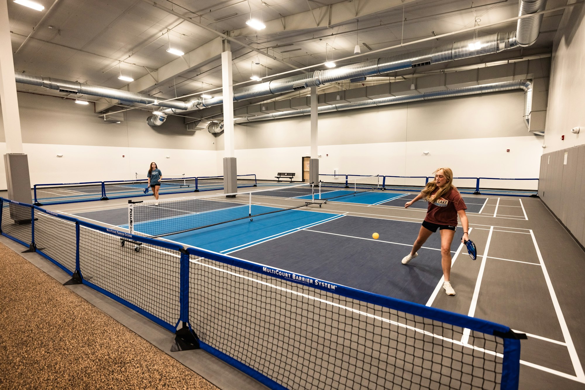 Two people playing pickleball indoors with blue courts and a viewing area in the background.