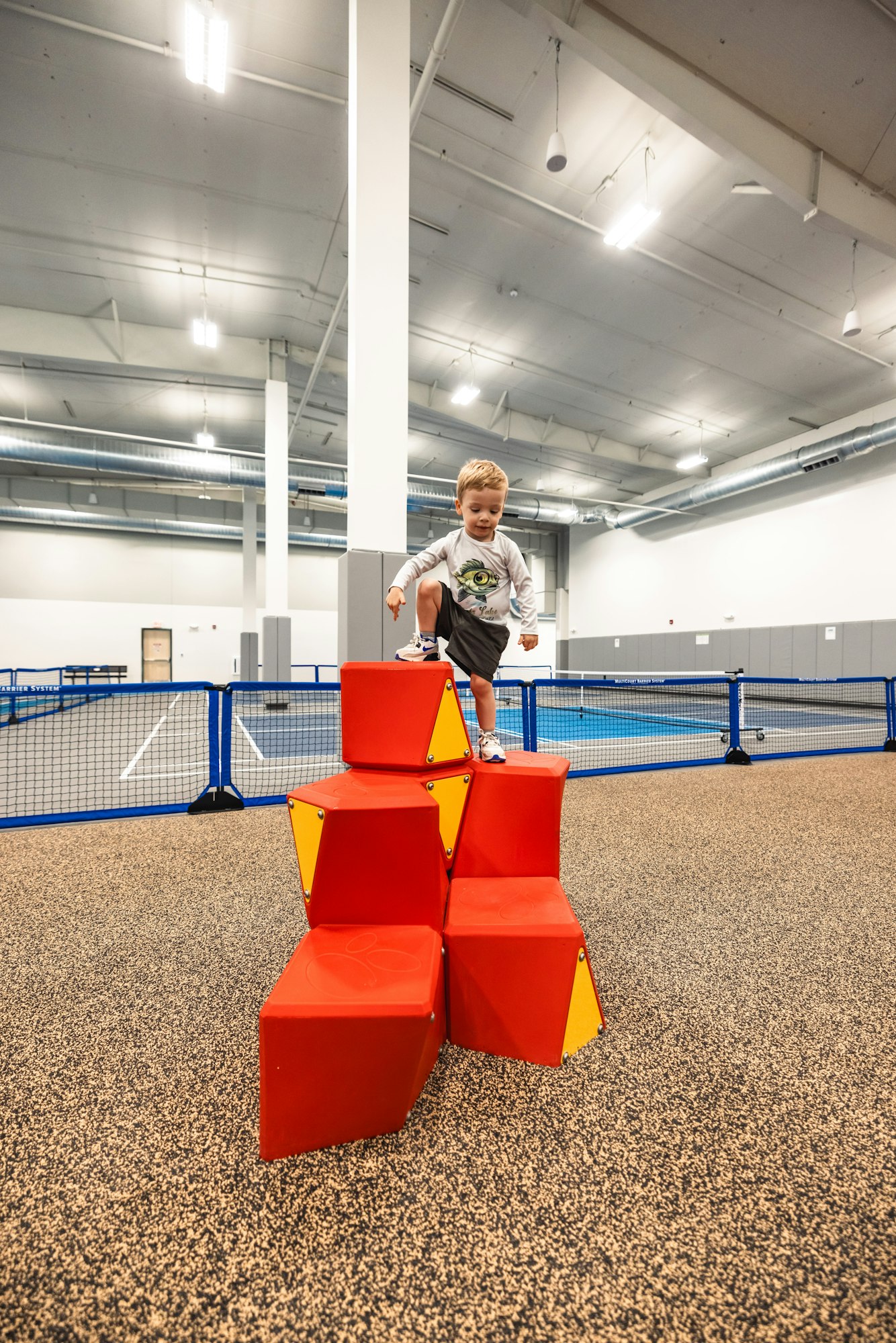 A child stands atop red foam blocks in an indoor play area with a blue net barrier in the background.