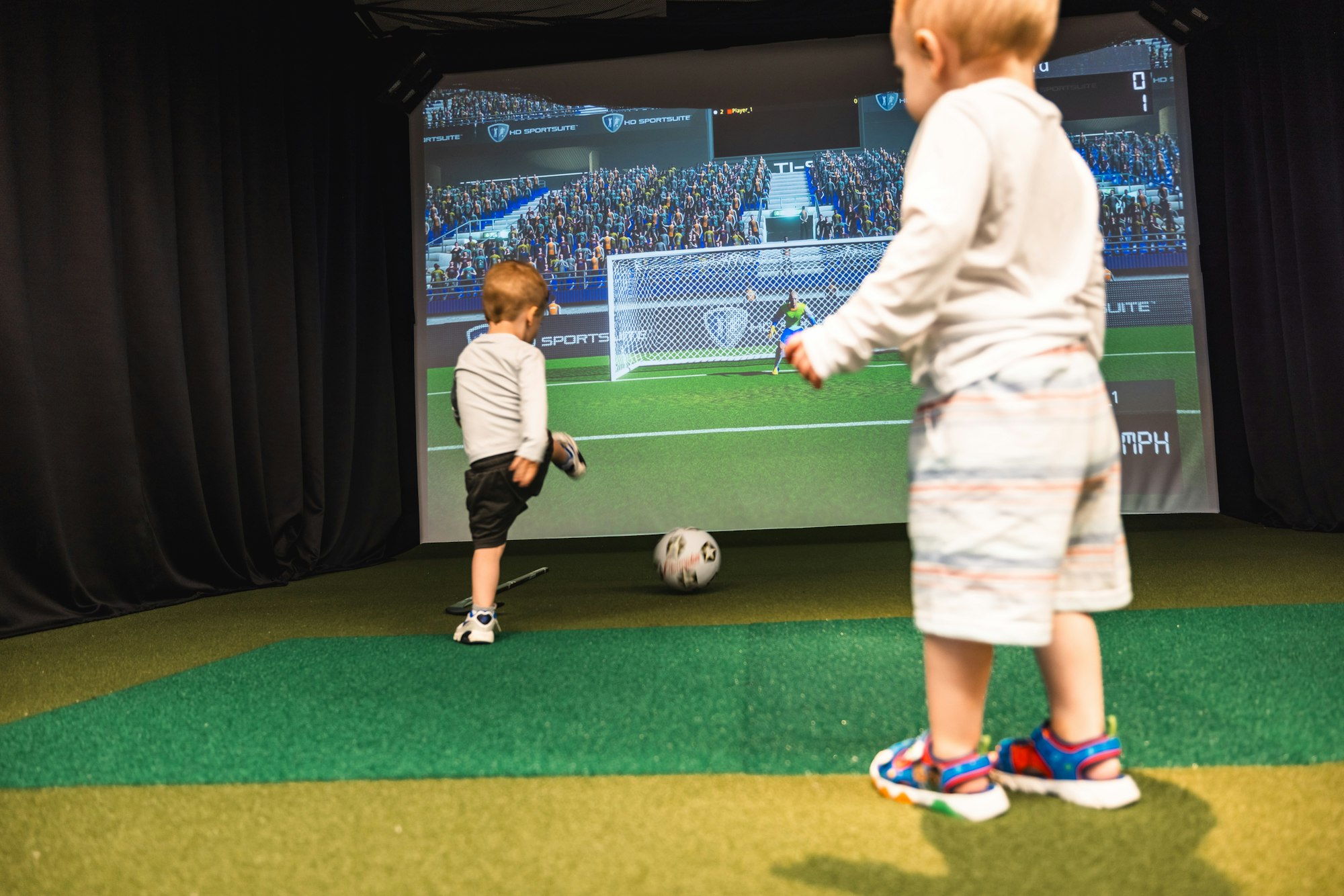 Two children playing with a soccer ball in front of a large screen simulating a soccer game.
