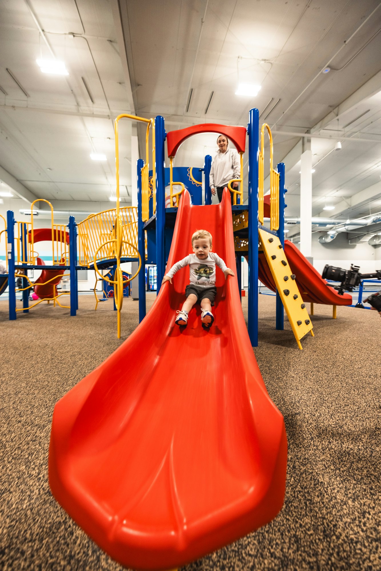 A child sliding down a red slide with another child at the top, in an indoor playground.