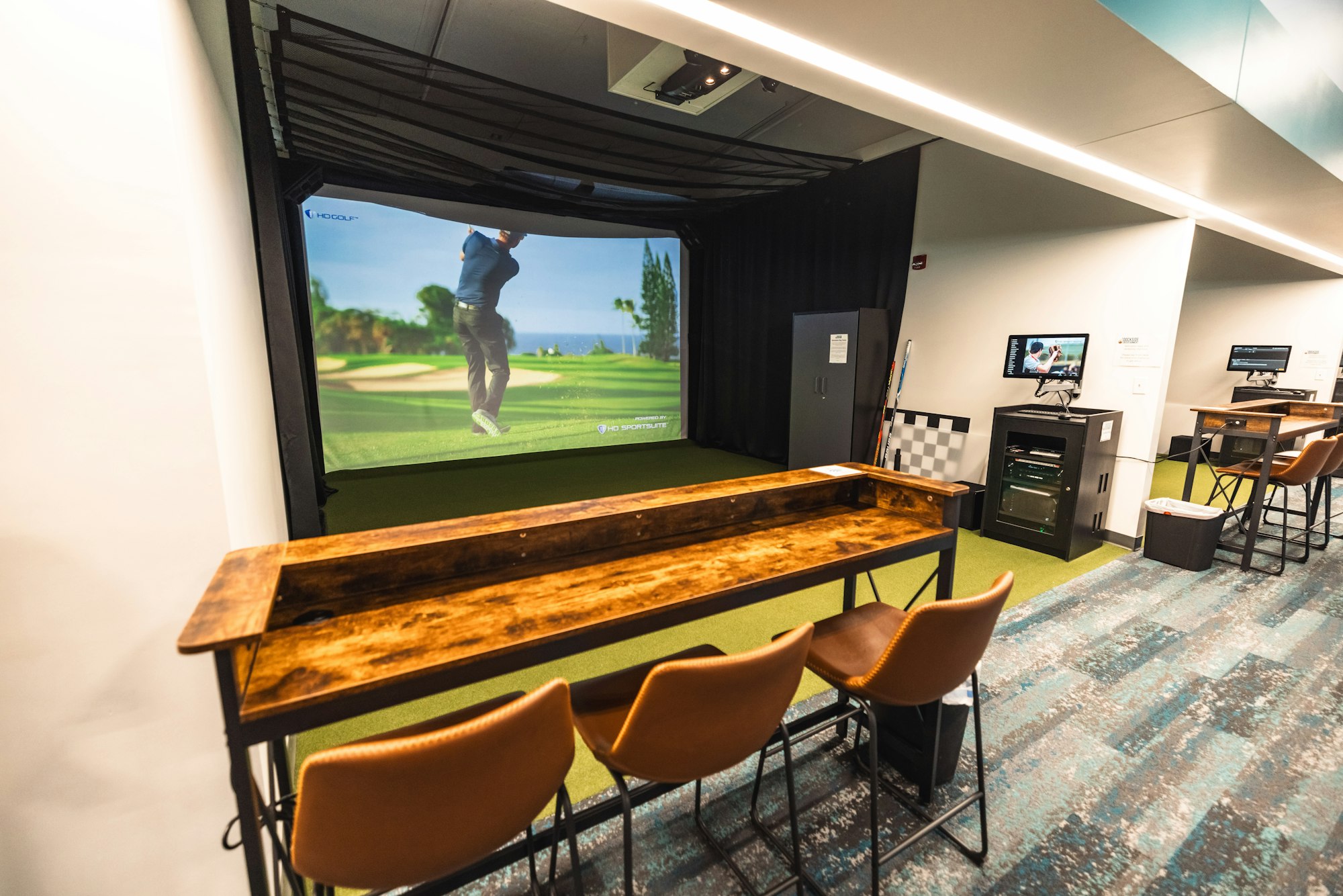 An indoor golf simulator room with a projected screen, seating area, and golf equipment.