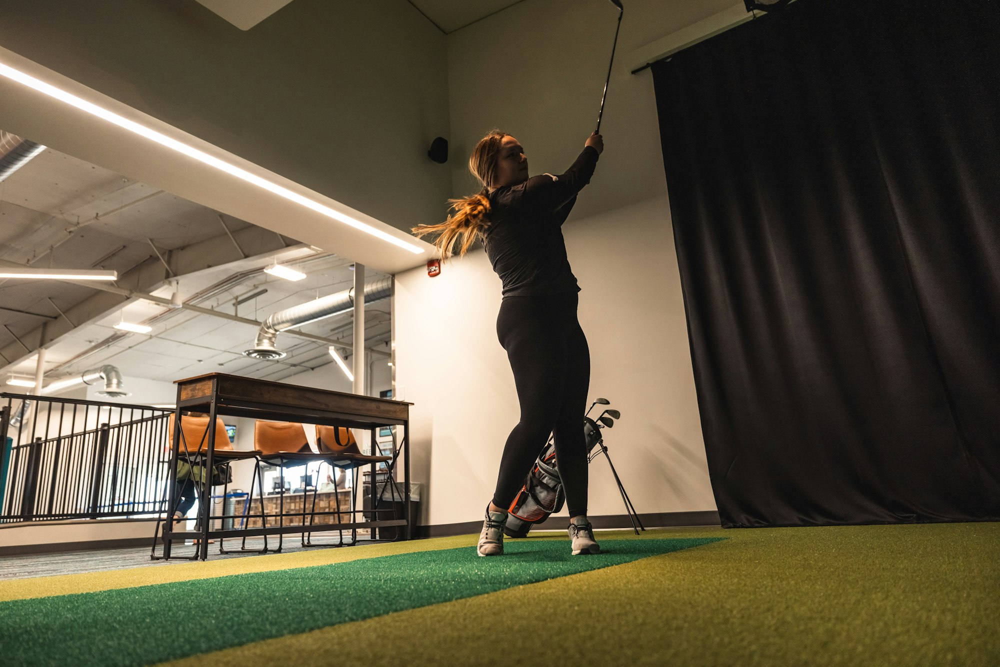 A woman practicing golf indoors, with clubs and artificial turf visible.