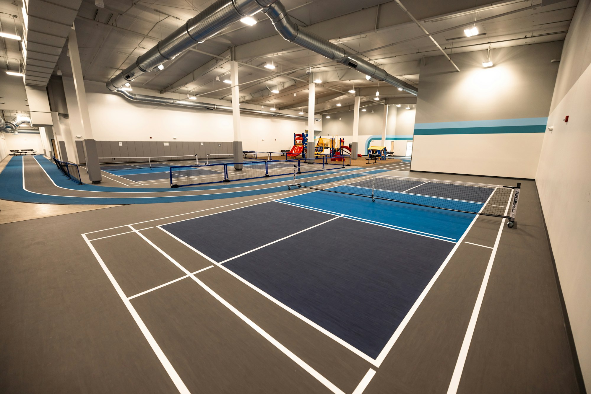 An indoor sports facility with multiple pickleball courts, bright lighting, and a running track along the edge.