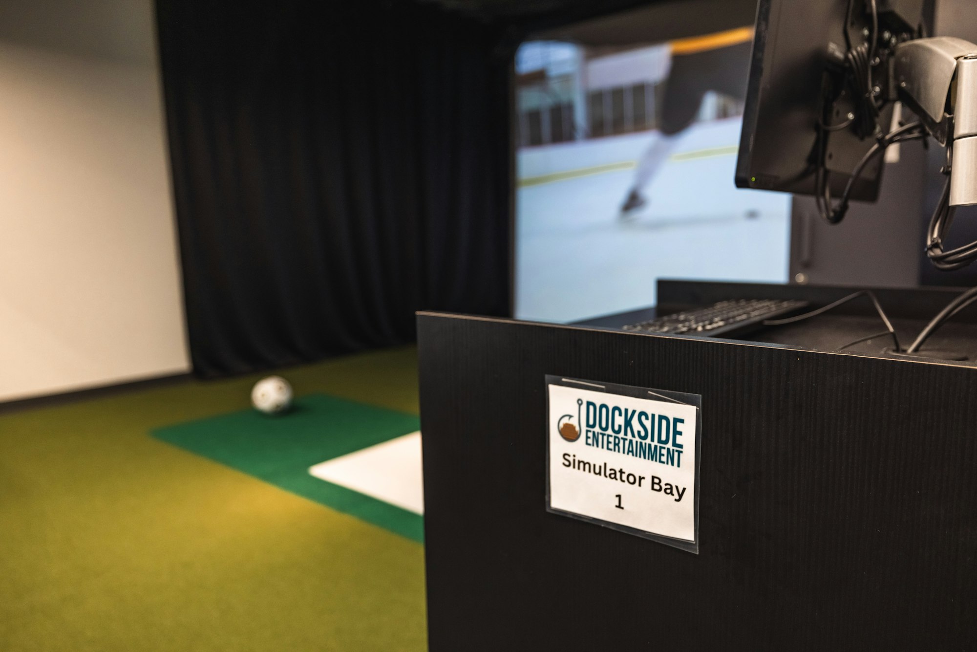 Golf simulator setup with screen, artificial turf, and signage indicating "Simulator Bay 1."