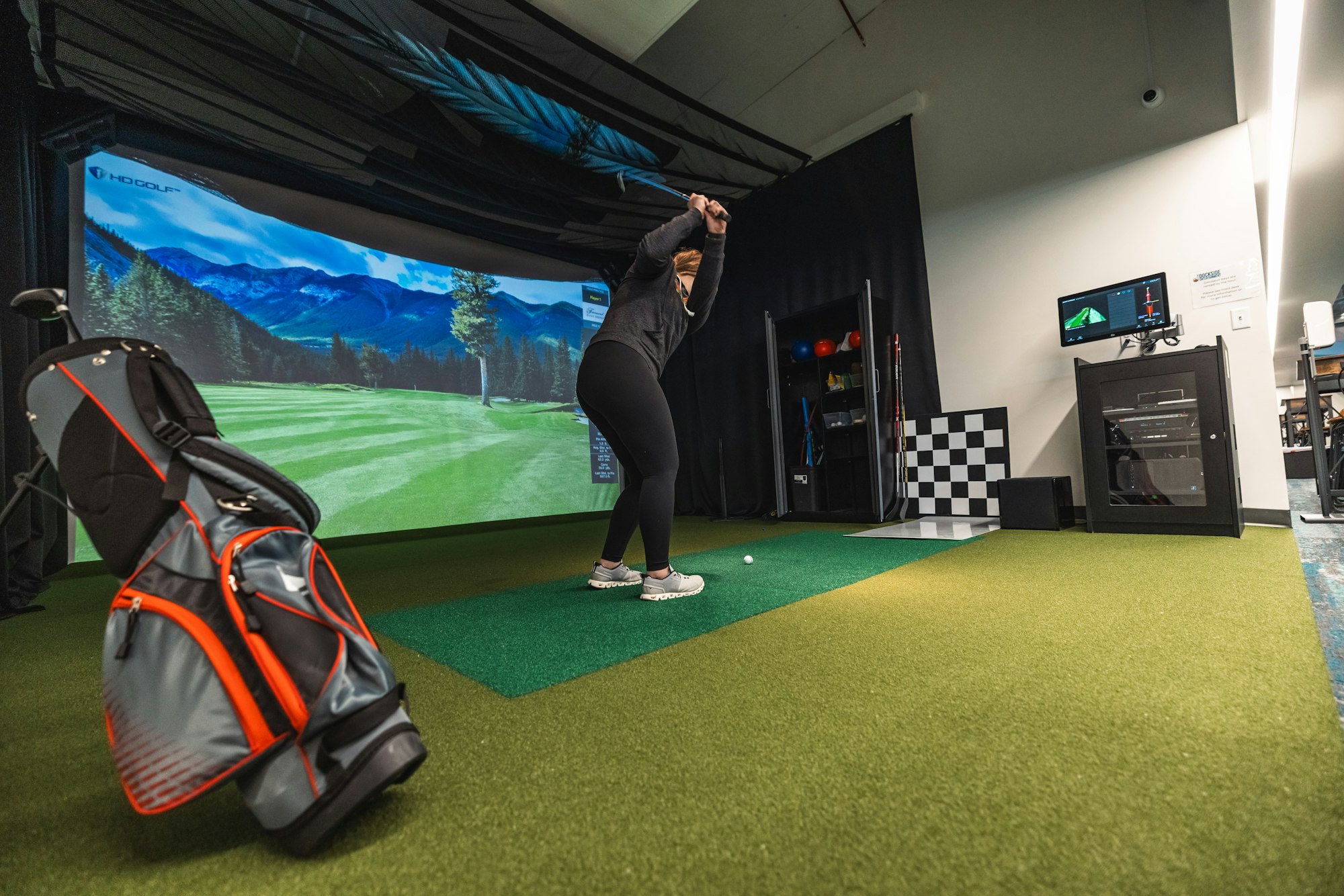 Person playing indoor golf simulator with a projected image of a golf course on the screen. Golf bag in the foreground.