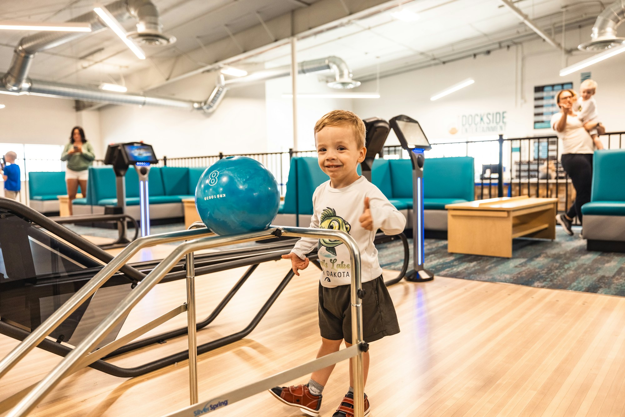 A young child in a bowling alley with a ball on the return ramp giving a thumbs-up. People in the background.