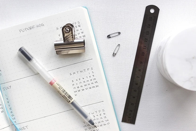 A planner with a pen, a binder clip, two paper clips, a ruler, and part of a white mug on a desk.