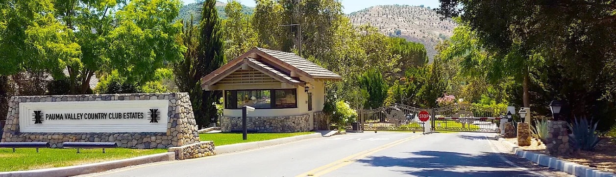 Entrance to "Pauma Valley Country Club Estates" with sign, guardhouse, trees, and mountain backdrop.
