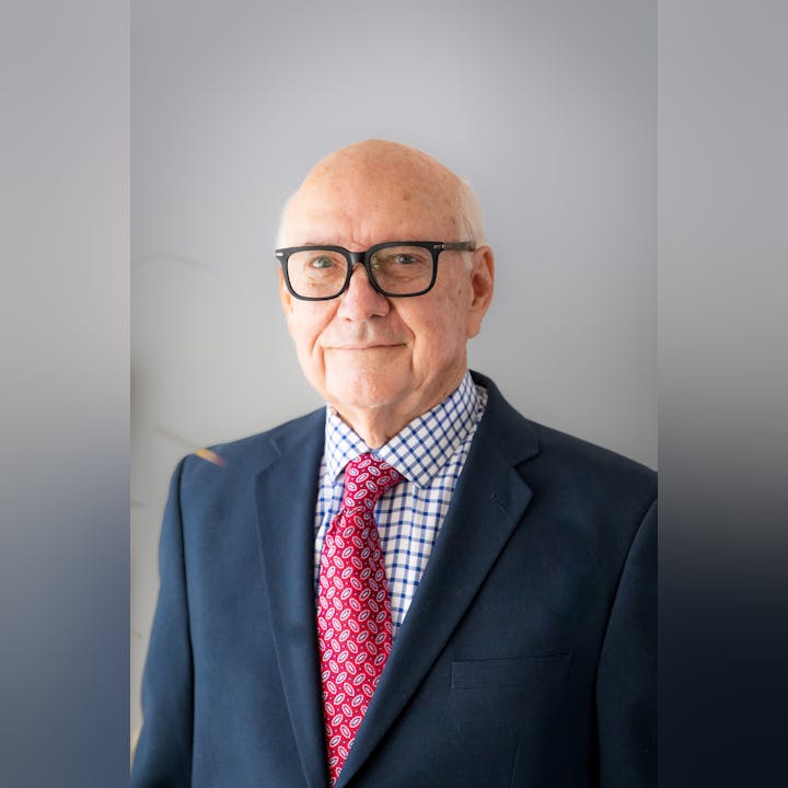 An older man wearing glasses, a suit, and a tie, stands in front of a plain background.