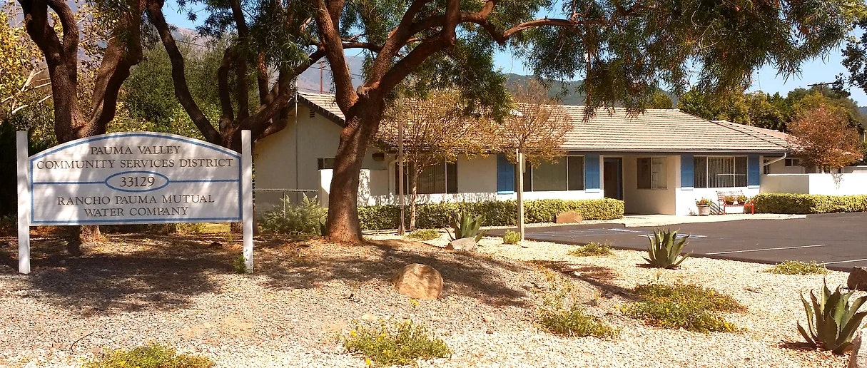 Single-story building with a sign for Pauma Valley Community Services District and Rancho Pauma Mutual Water Company, surrounded by trees.