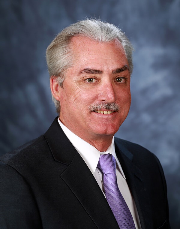 The image shows a man with silver hair and a mustache, wearing a suit and tie, smiling against a blue background.