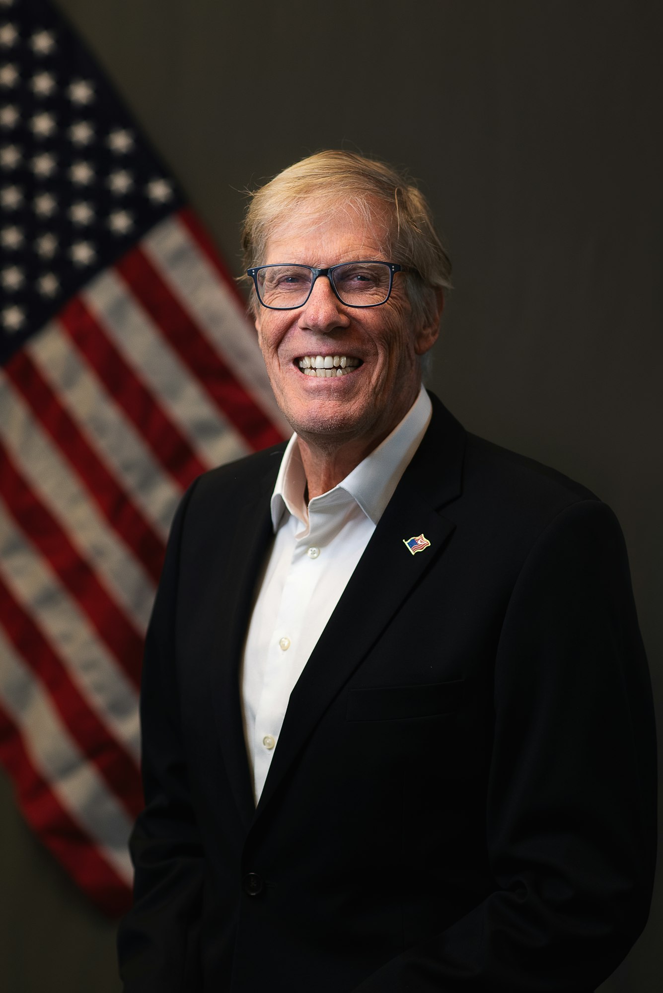 A man in a suit with glasses smiles in front of a U.S. flag.