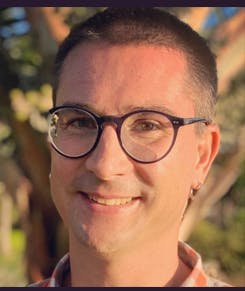 A person wearing glasses, smiling outdoors with a blurred background of trees.