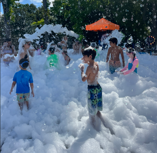 Kids are playing joyfully in a large foamy bubble area on a sunny day, surrounded by trees and a tent.