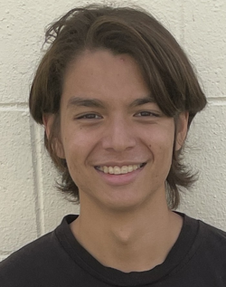 A person with medium-length hair, wearing a black shirt, smiling against a light-colored textured wall.