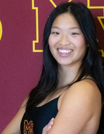 A person smiling with long dark hair, wearing a black sleeveless top against a maroon and yellow background.