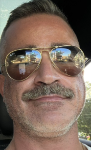 A person with sunglasses and a mustache, outdoors with a reflection of the surroundings visible in the glasses.