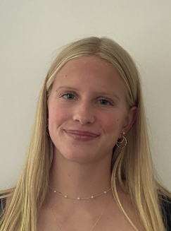A person with long blonde hair and earrings, smiling slightly, against a plain background.
