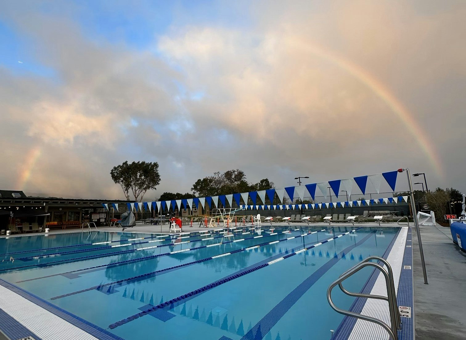 Outdoor swimming pool with lanes, bunting flags, and a rainbow in the sky.