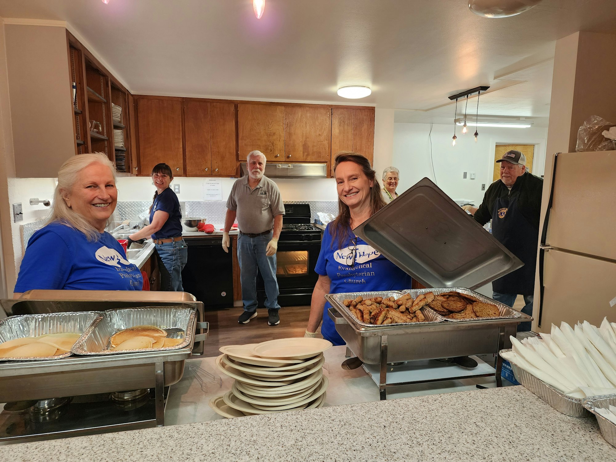 Smiling people serving food at a community or church kitchen.