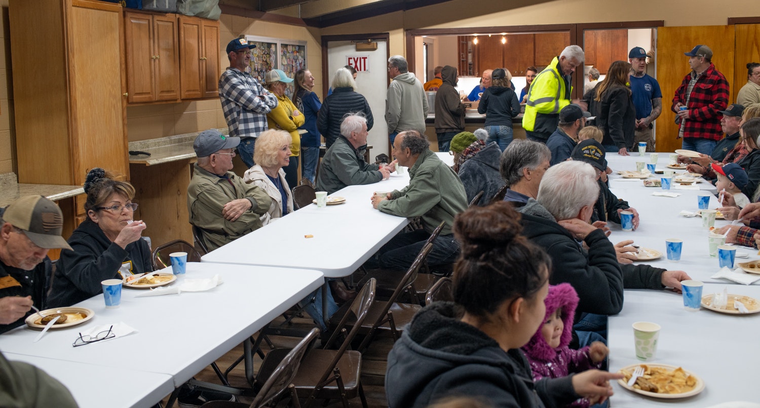 A group of people gathered, eating and socializing at long tables in a community hall or similar venue.