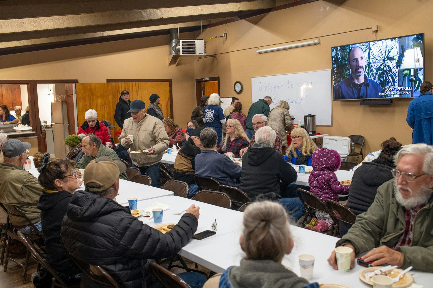 A group of people gathered in a room with tables, eating and conversing, with a TV screen in the background.