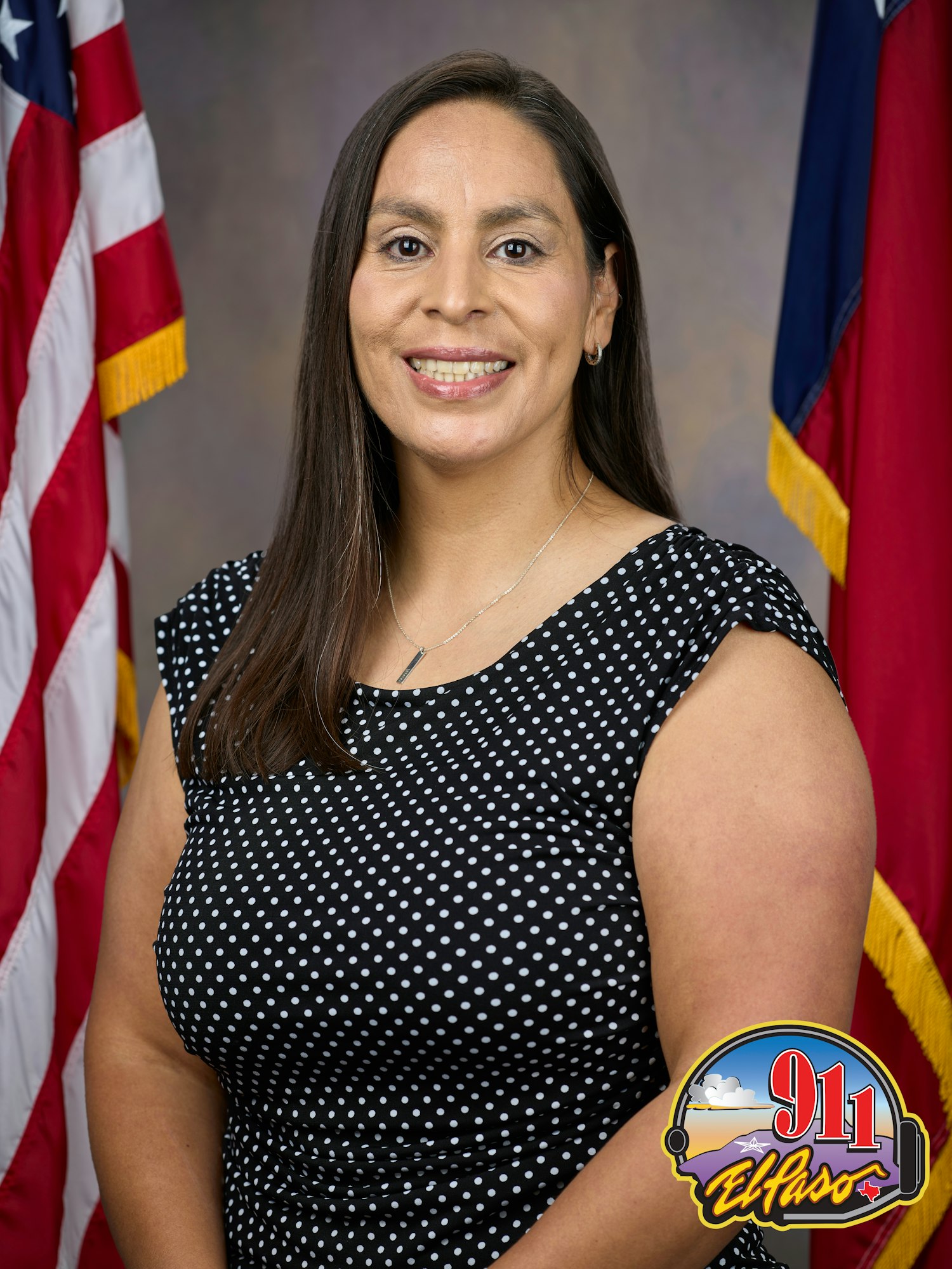 A woman standing in front of the US and Texas flags, wearing a polka dot top and a badge that says "911 El Paso".