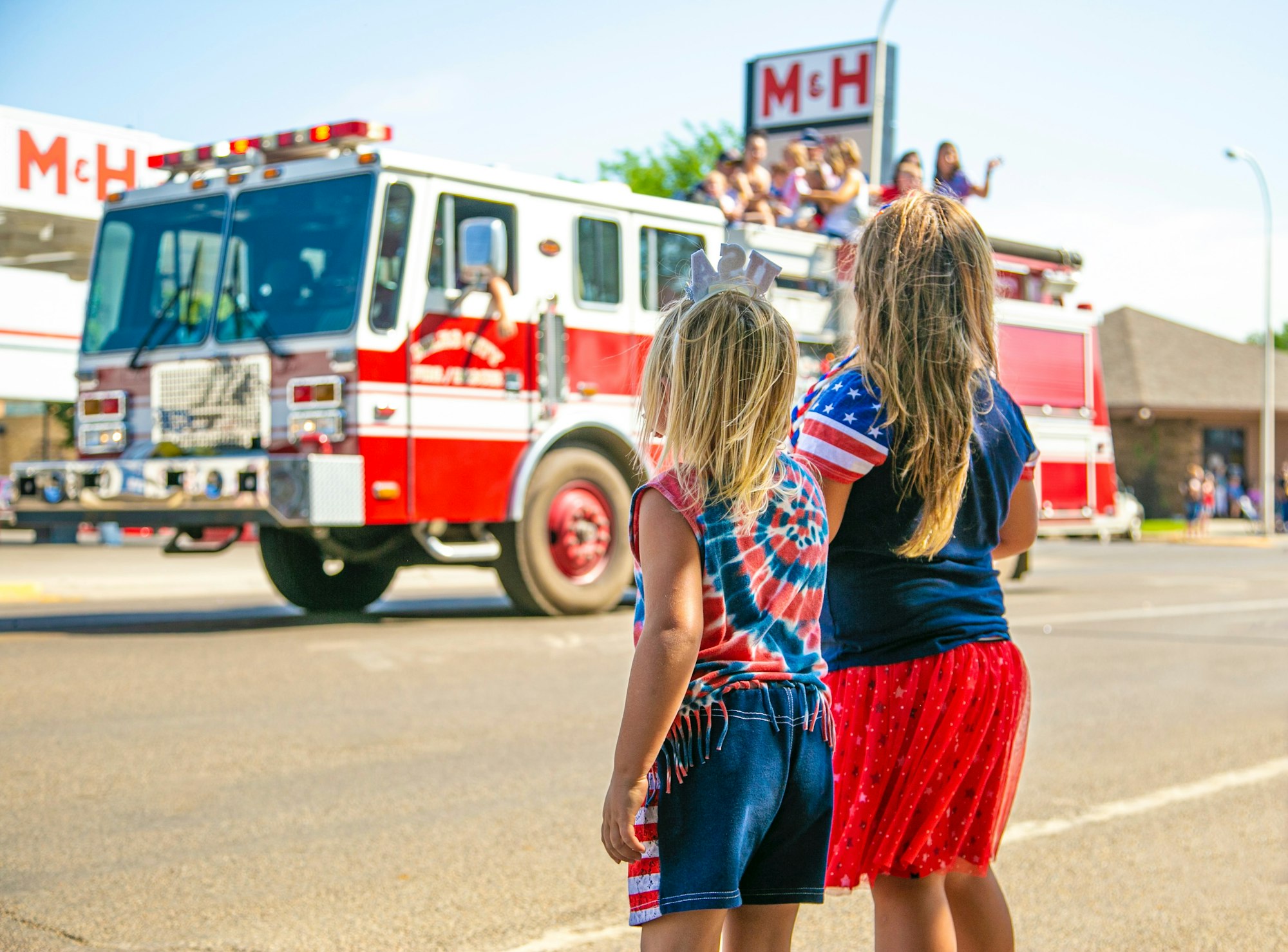 Two children watch a fire truck during a parade, dressed in festive, patriotic clothing under a bright blue sky.